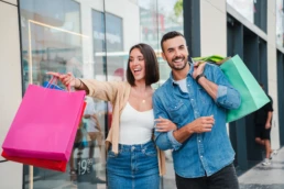 Um casal, composto por um homem e uma mulher, com sacolas de compras, aponta e sorri para uma vitrine no shopping enquanto decide sobre a compra de uma oferta, possivelmente um presente.