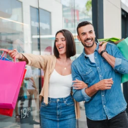 Um casal, composto por um homem e uma mulher, com sacolas de compras, aponta e sorri para uma vitrine no shopping enquanto decide sobre a compra de uma oferta, possivelmente um presente.