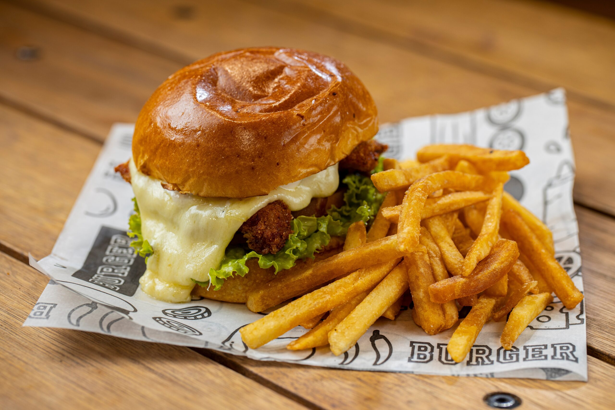 Cheeseburger com frango crocante, queijo derretido e alface, acompanhado de batatas fritas douradas em uma bandeja de papel.