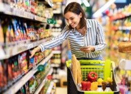 Mulher jovem sorrindo segura um smartphone enquanto pega alimentos saudáveis de uma prateleira no supermercado.