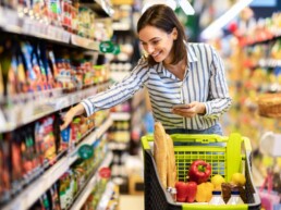 Mulher jovem sorrindo segura um smartphone enquanto pega alimentos saudáveis de uma prateleira no supermercado.