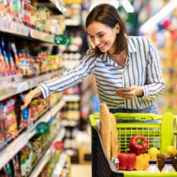 Mulher jovem sorrindo segura um smartphone enquanto pega alimentos saudáveis de uma prateleira no supermercado.