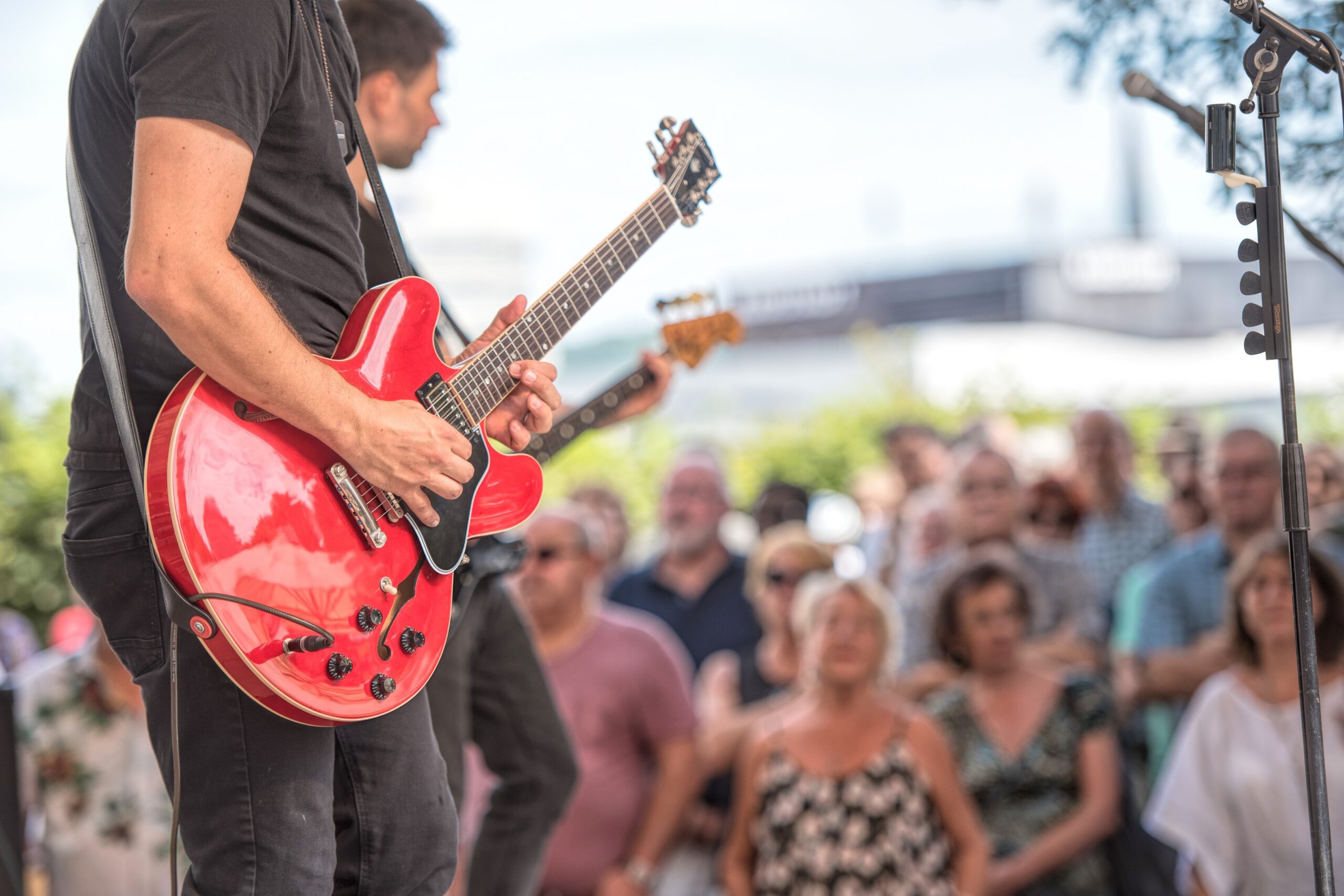  Foto de um guitarrista tocando guitarra vermelha durante um show ao vivo, com foco no instrumento e a multidão assistindo ao fundo.