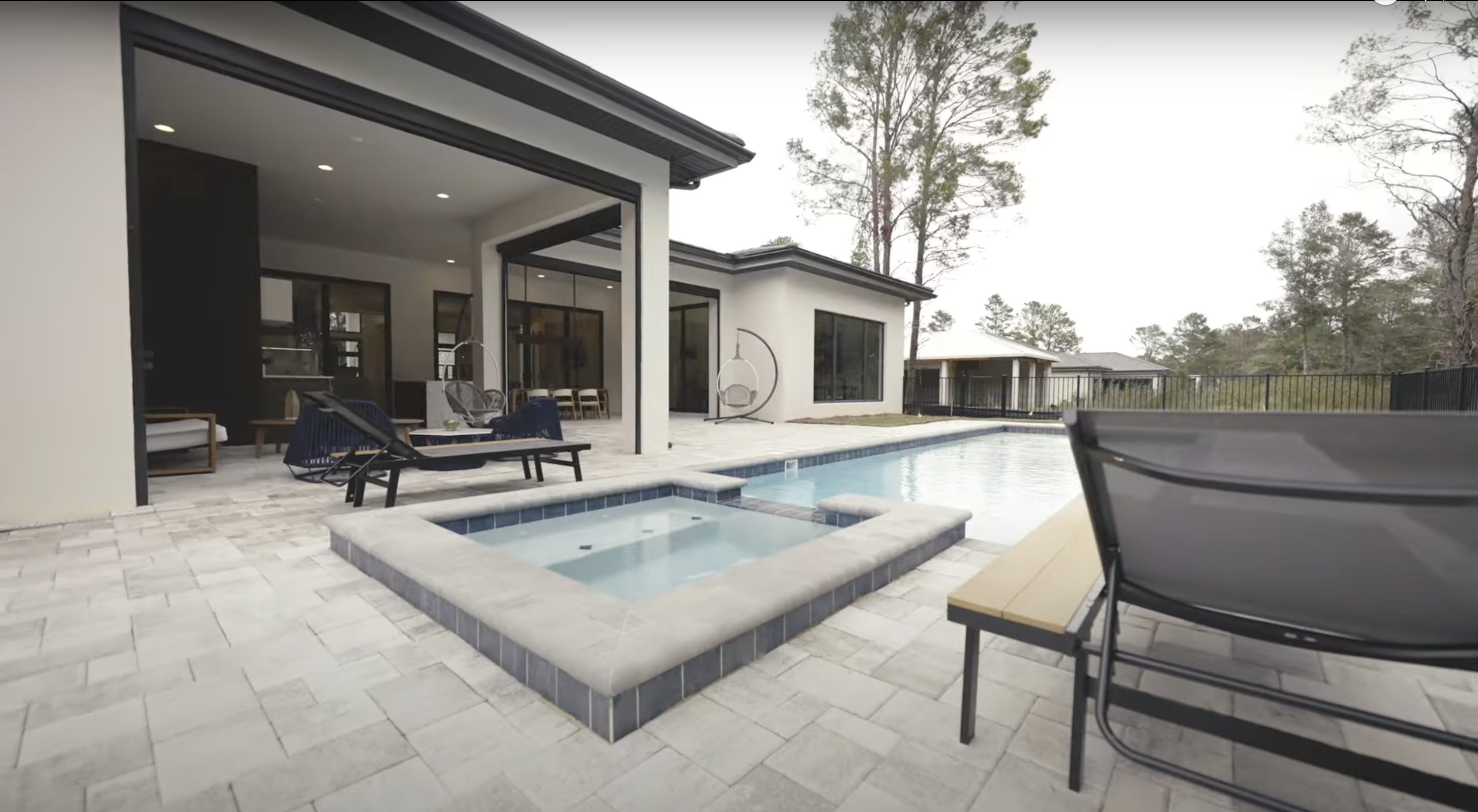 Vista da área externa de uma casa moderna com piscina, jacuzzi e cadeiras de descanso em um pátio cercado por ladrilhos claros e árvores ao fundo.