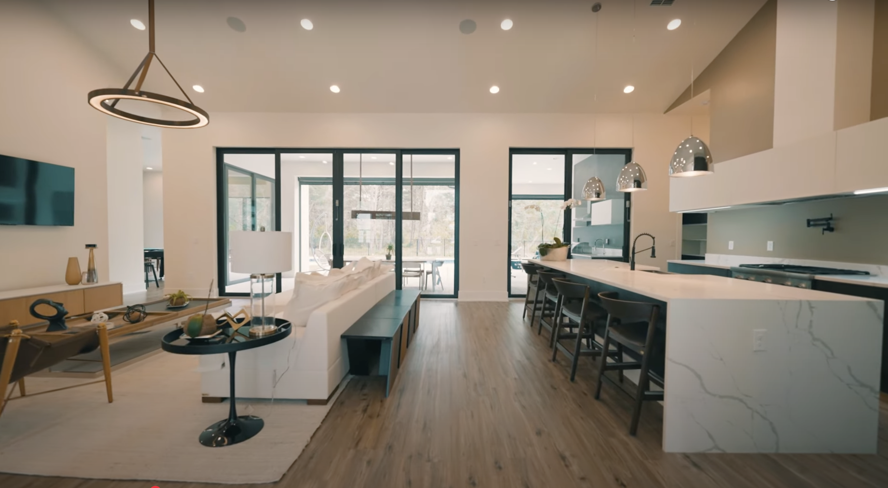 Sala de estar moderna com sofá branco, mesa de centro de madeira, cozinha com bancada de mármore e luminárias suspensas.