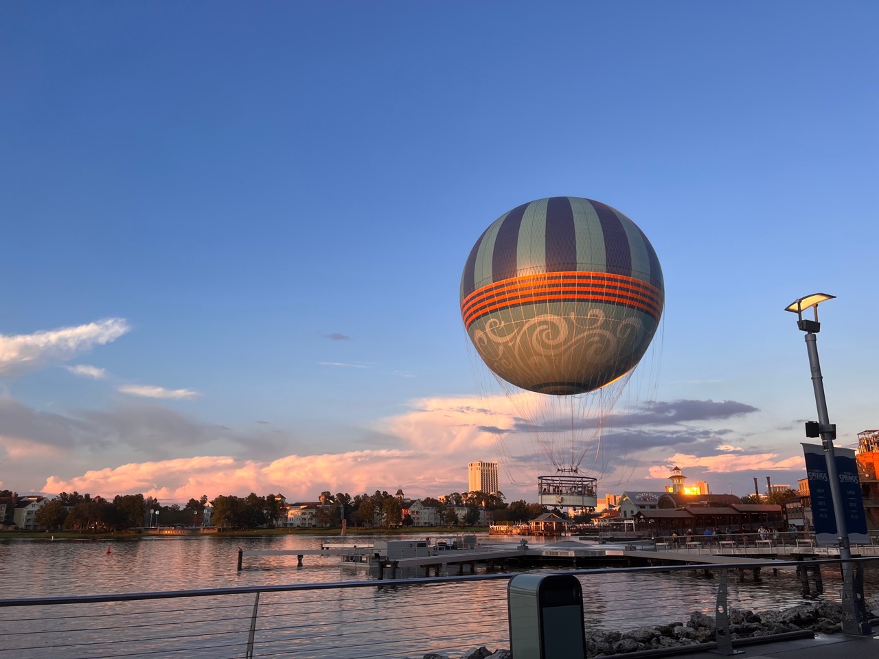 Balão de ar quente azul e laranja, com detalhes decorativos, pairando sobre um lago ao entardecer, com céu misturado de azul e dourado ao fundo.