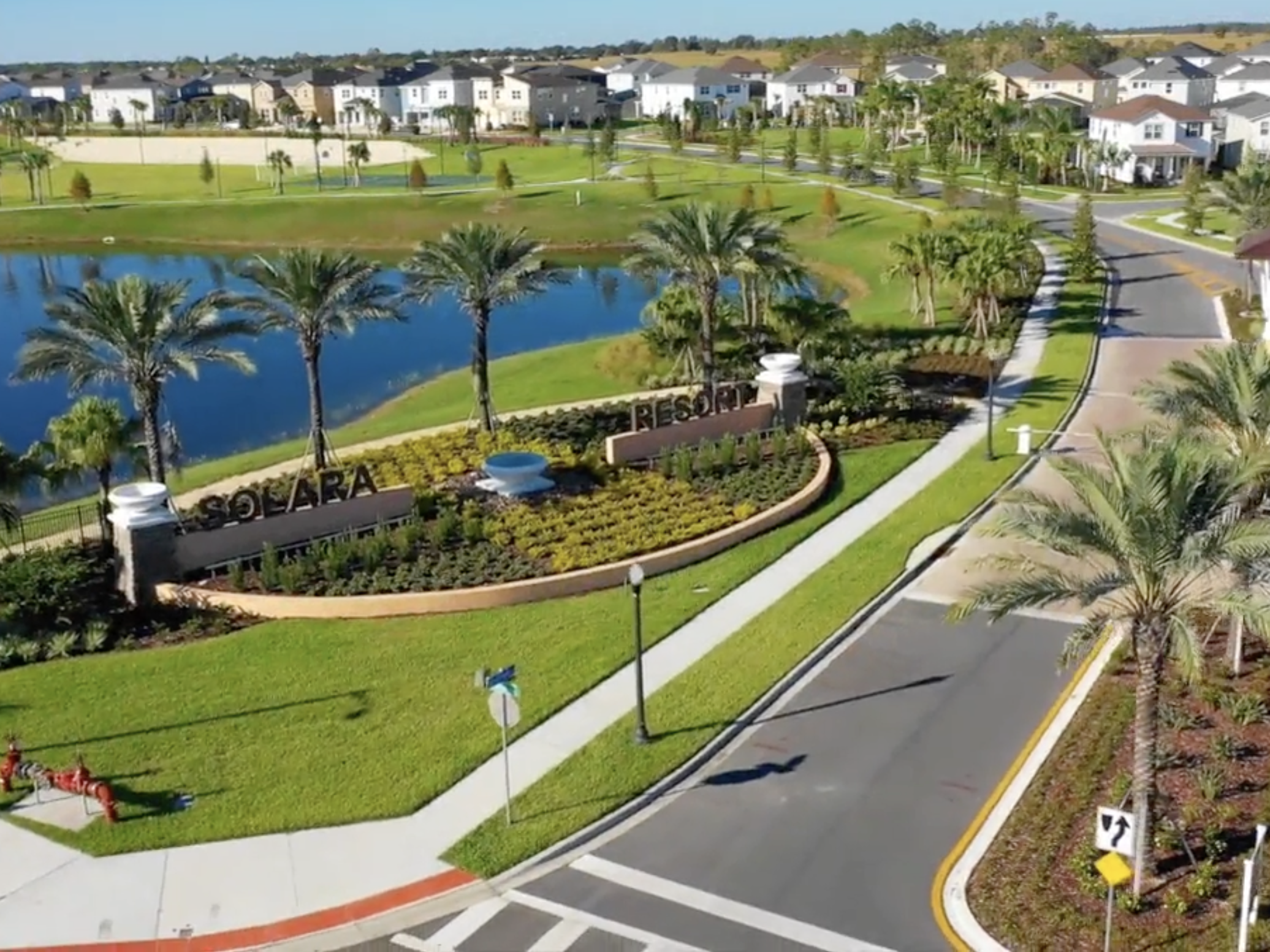 Entrada do condomínio Solara Resort com paisagismo, palmeiras, lago ao fundo e casas residenciais ao redor.