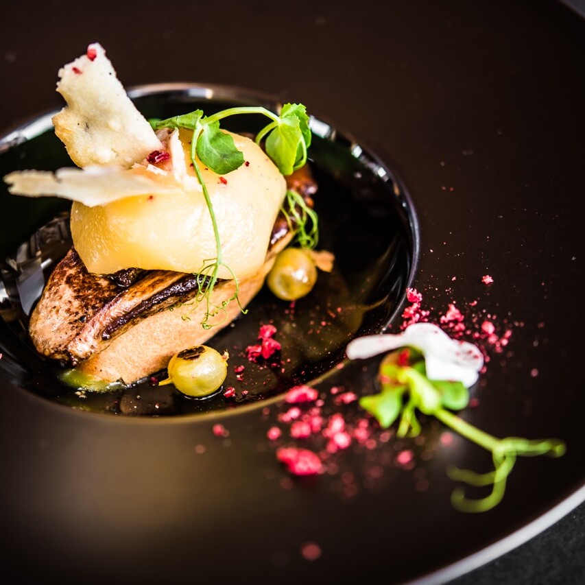 Prato gourmet com foie gras grelhado, pera caramelizada, microverdes e decoração artística em um prato escuro.