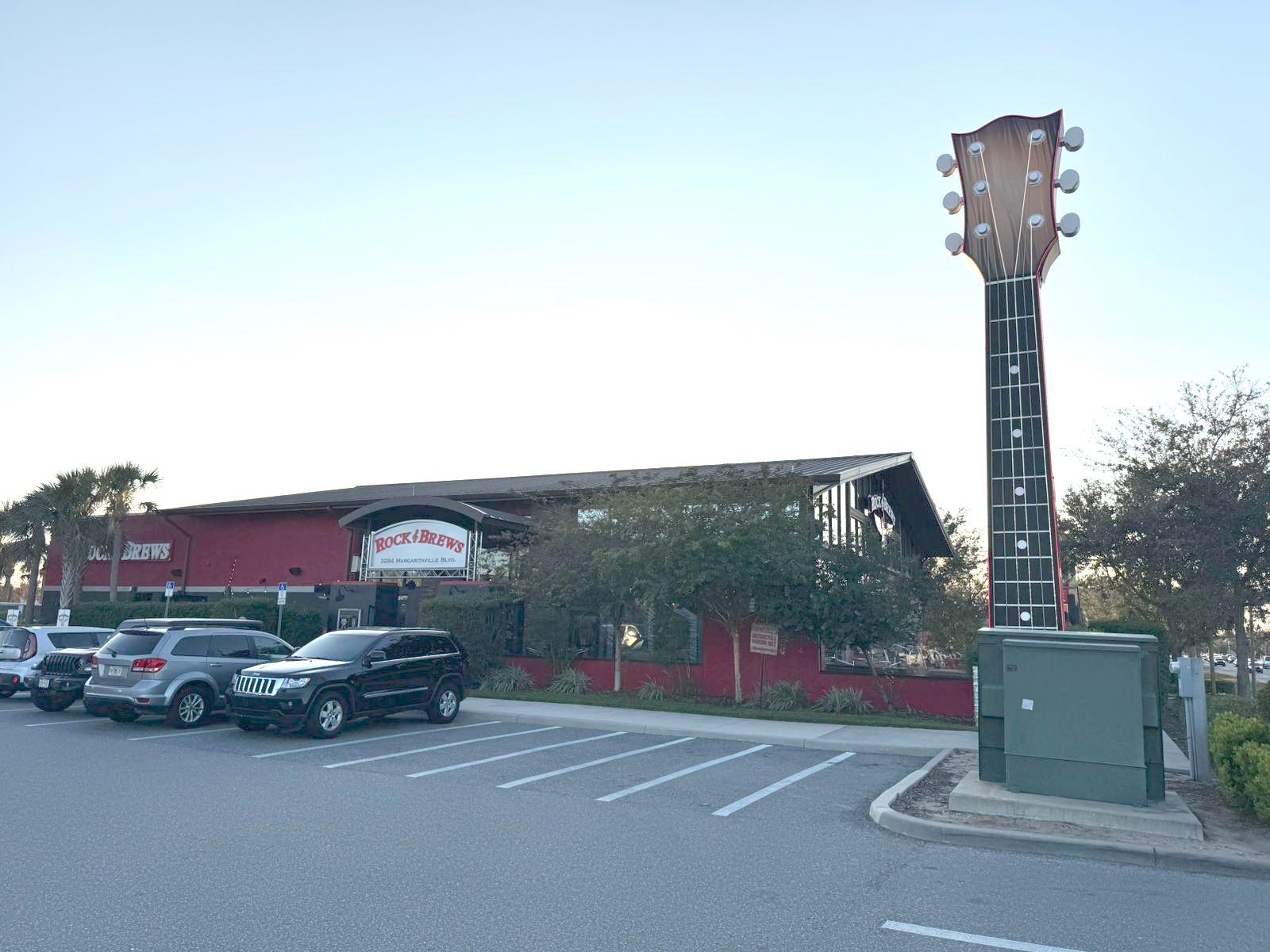 Restaurante Rock & Brews com fachada vermelha e uma grande escultura de braço de guitarra ao lado, cercado por árvores e um estacionamento com veículos.