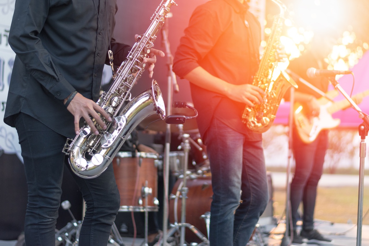 Duas pessoas tocando saxofone em um palco ao ar livre, acompanhadas de uma banda, com iluminação natural e reflexos do pôr do sol ao fundo