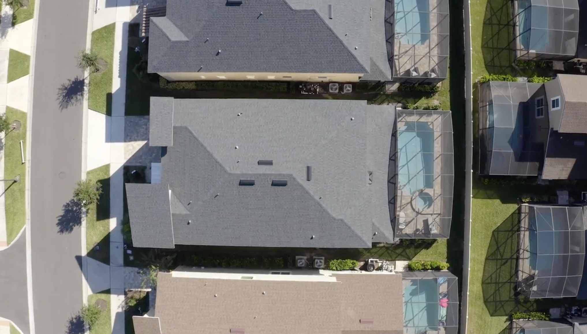 Vista aérea de telhados de casas em um bairro residencial, com piscinas cobertas por telas e gramados ao redor.