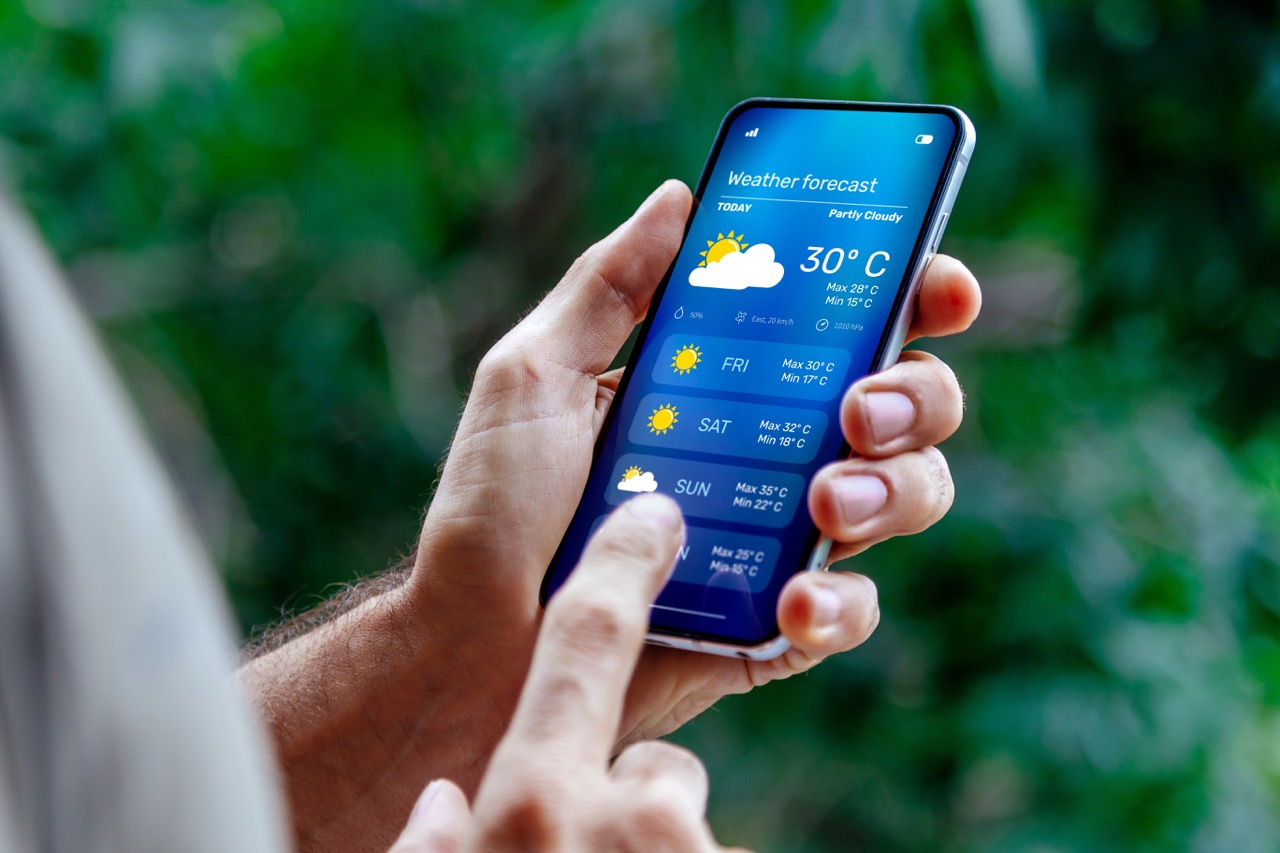 Mão de uma pessoa segurando um smartphone com a tela exibindo um aplicativo de previsão do tempo, incluindo temperaturas e condições climáticas.